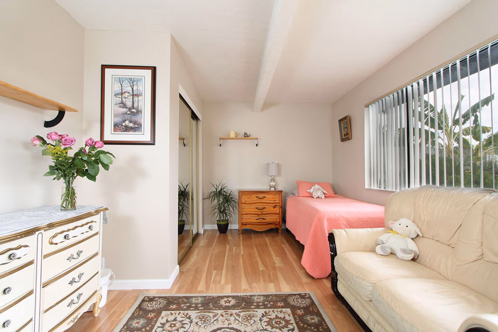 Bright single-occupancy bedroom with a pink-covered twin bed, wooden nightstand and dresser, a cream loveseat, and a large window with vertical blinds.