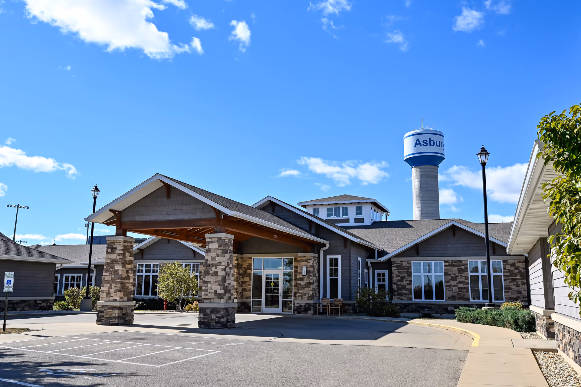 Exterior front entrance of a single-story senior living building with a covered porte-cochère and a water tower in the background.