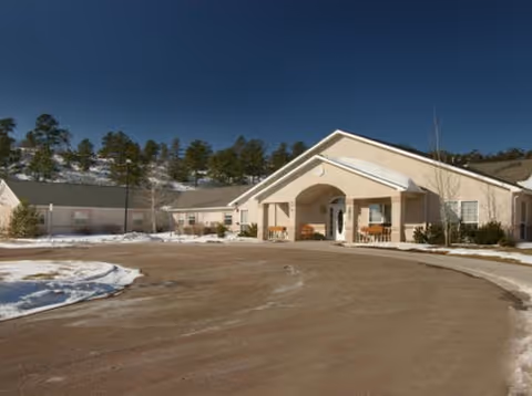 Entrance of a single-story senior living facility with a covered portico, curved driveway, and patches of snow under a clear blue sky.