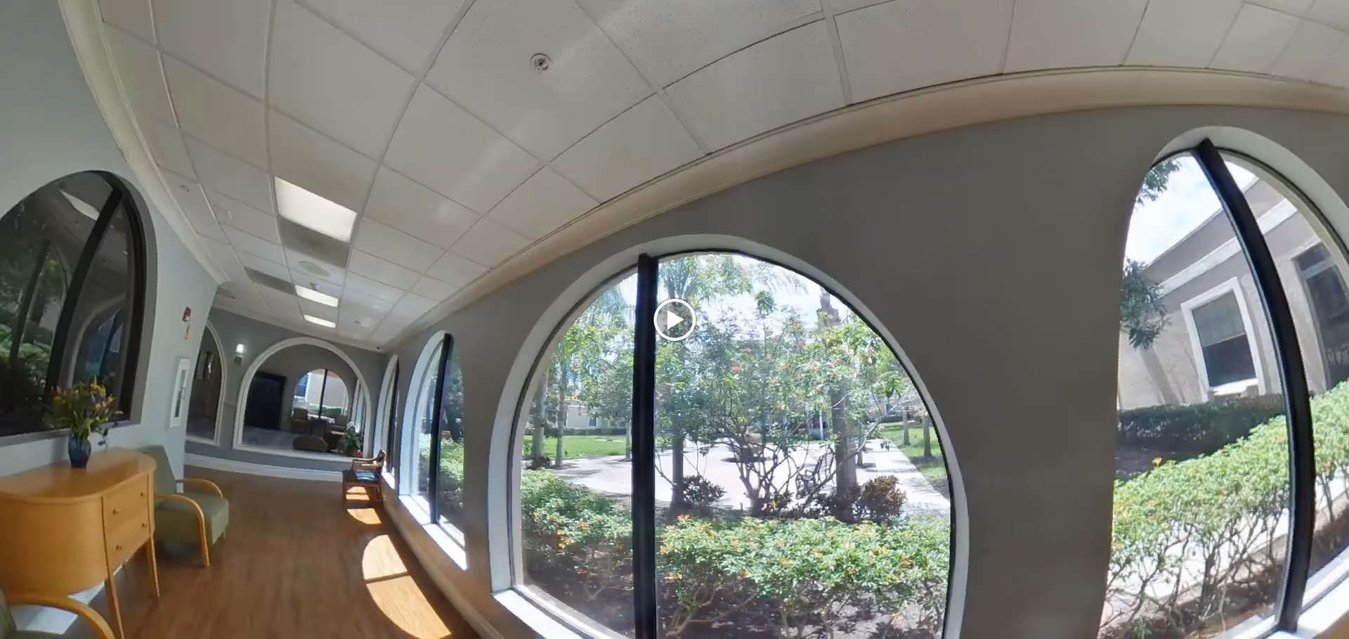 A bright indoor hallway with large arched windows on the right side showing a view of green bushes and trees outside. The hallway has wooden flooring, light gray walls, and a white ceiling with recessed lighting. There is a small wooden cabinet with a vase of flowers and a green chair on the left side near the windows.