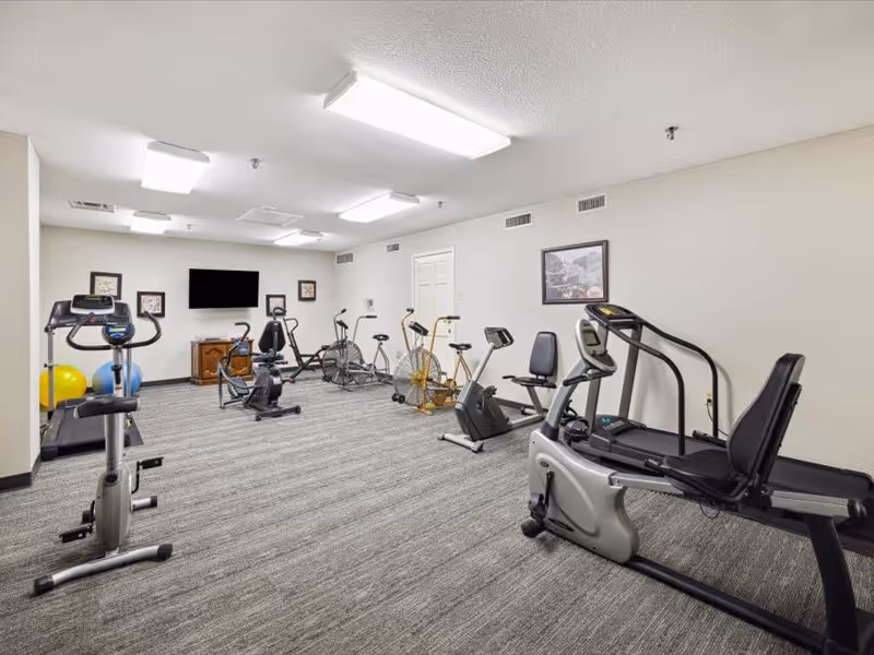 A fitness room with various exercise equipment including stationary bikes, an elliptical machine, and exercise balls. The room has gray carpeted flooring, white walls, fluorescent ceiling lights, and a wall-mounted TV. There are framed pictures on the walls and a wooden cabinet beneath the TV.