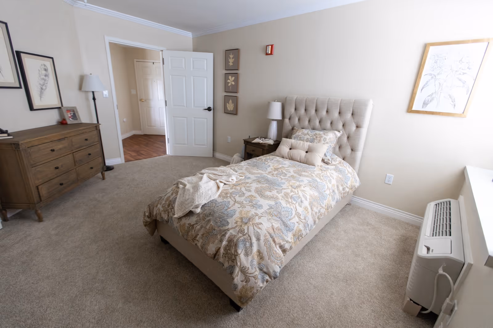 A cozy bedroom in a senior living facility featuring a single bed with a tufted headboard and floral bedding. The room has beige walls, carpeted floor, a wooden dresser with framed artwork above it, a floor lamp, and a nightstand with a lamp. There is a white door open to a hallway with wooden flooring and a wall-mounted air conditioning unit below a window.