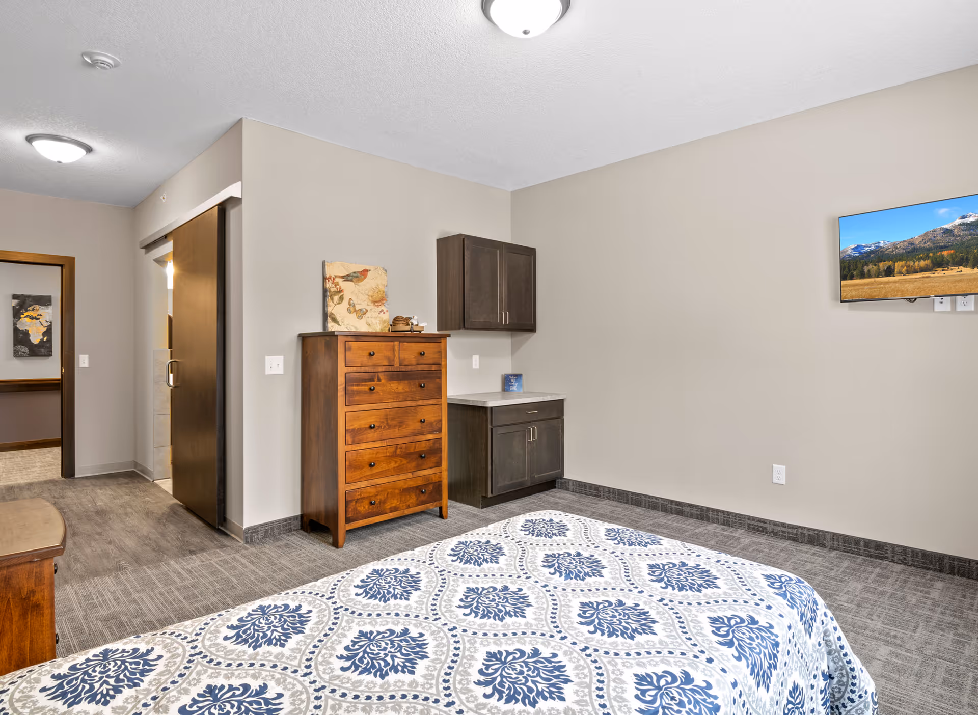 A bedroom in a senior living facility with a bed covered in a blue and white patterned bedspread. The room features a wooden chest of drawers, a small cabinet with a countertop, a wall-mounted flat-screen TV displaying a mountain landscape, and a sliding wooden door leading to another area. The walls are painted beige and the floor is carpeted.