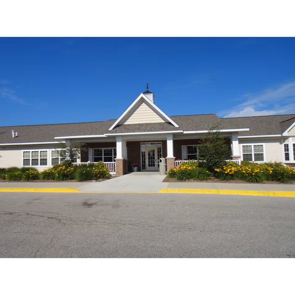 Front exterior view of a single-story assisted living facility building with a peaked roof entrance, white columns, brick accents, and surrounding landscaping with yellow flowers and green shrubs under a clear blue sky.