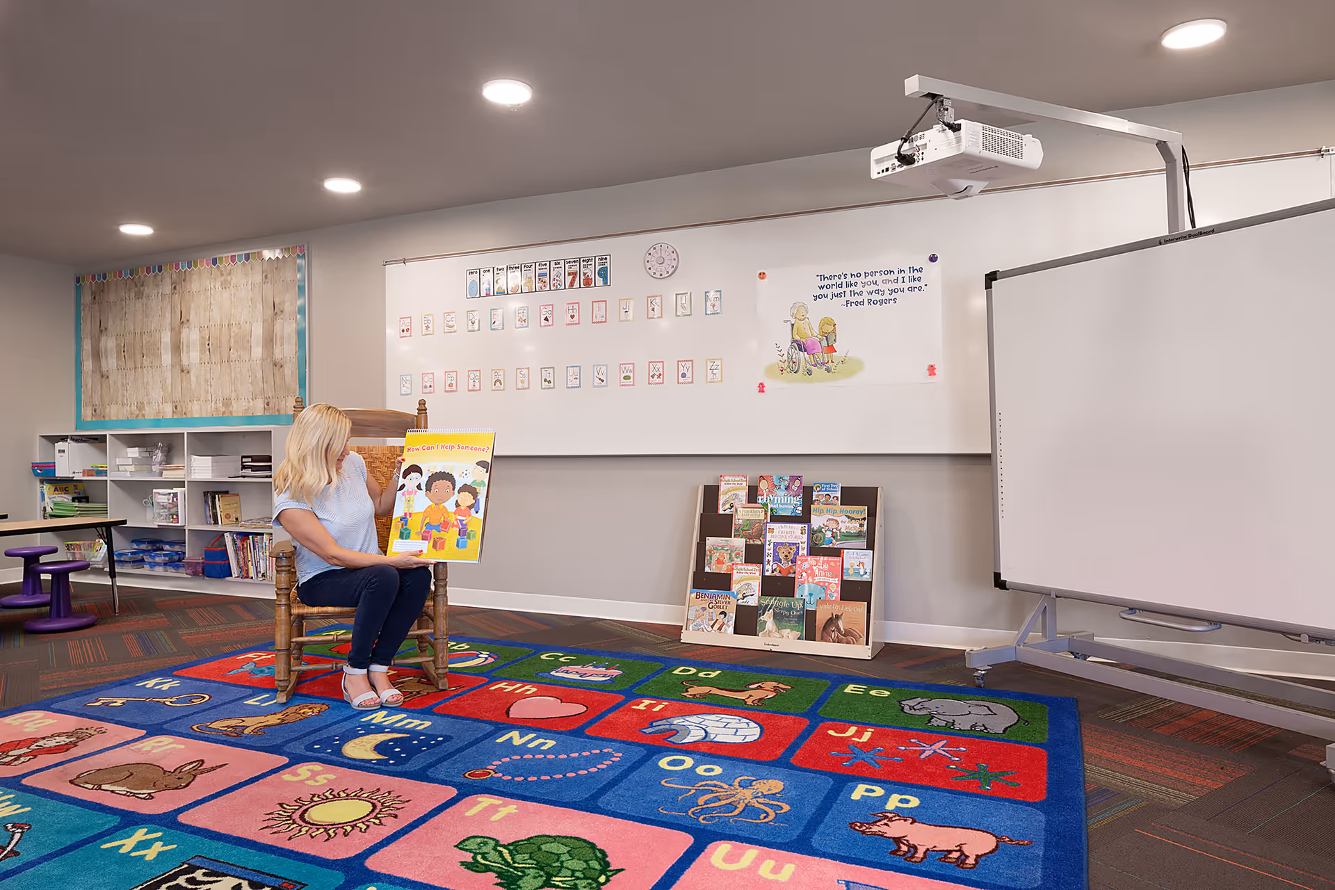 A woman with blonde hair sits on a wooden chair on a colorful alphabet rug in a classroom-like setting. She is holding up a children's book titled 'How Can I Help Someone?'. The room has a whiteboard with alphabet cards and a Fred Rogers quote, a projector mounted on the ceiling, and shelves with books and educational materials.