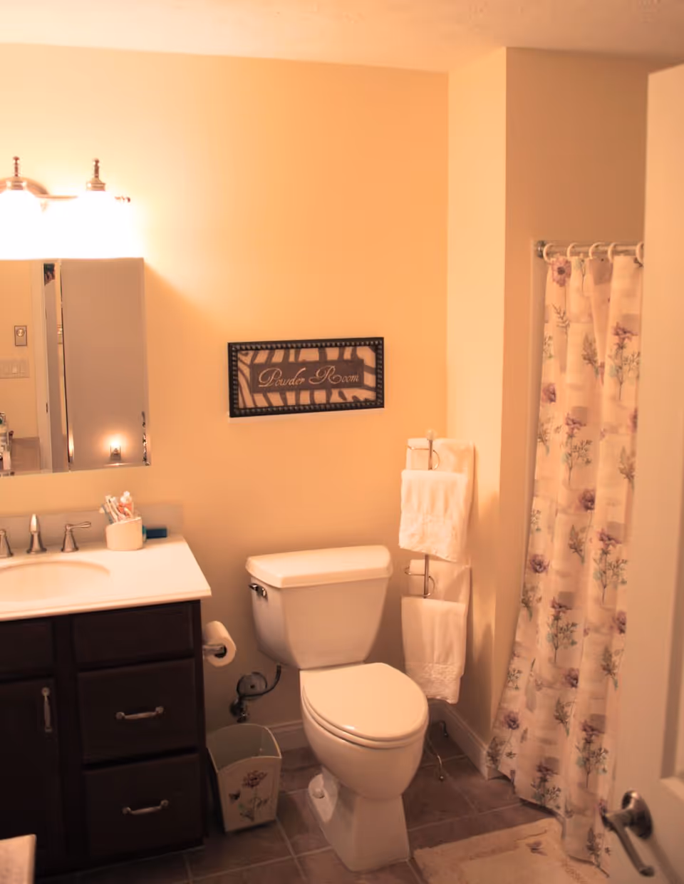 Small bathroom with a toilet, sink vanity and mirror, towel rack, and a floral shower curtain.