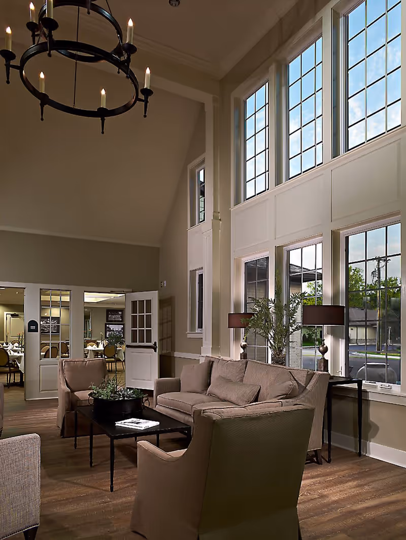 Two-story communal living room with large grid windows, beige sofa and armchairs arranged around a coffee table, lamps, and a chandelier overhead.