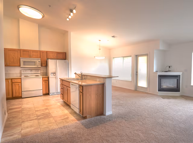 Open-plan interior with a kitchen island and oak cabinets opening to a carpeted living area with a fireplace and exterior door.