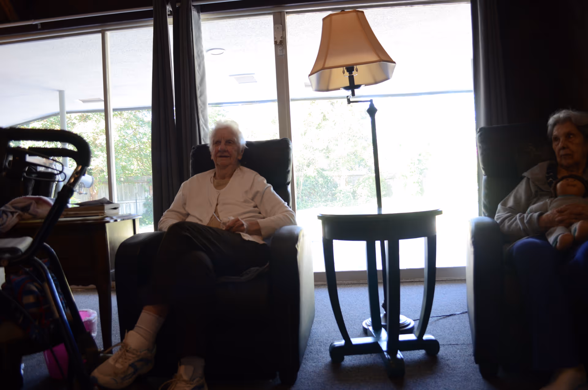 Two elderly women sitting in armchairs in a living room with a large window and a floor lamp on a small table between them. One woman is holding a doll, and there is a walker and a side table with items on it to the left.