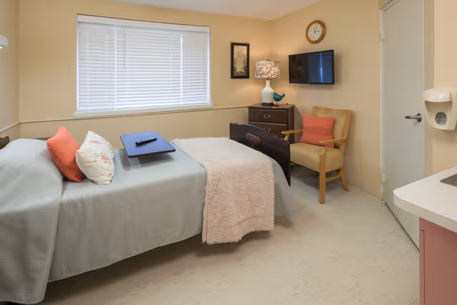 A small, neatly arranged bedroom in a senior living facility with a single bed covered in a light blue bedspread and a beige throw blanket. The bed has two pillows, one white with a floral pattern and one orange. A dark wooden tray with a remote control rests on the bed. Next to the bed is a wooden armchair with an orange cushion. A small dark wooden dresser with a decorative lamp and a blue bird figurine sits against the wall. Above the dresser is a wall-mounted flat-screen TV and a round clock. The room has beige walls, a window with white blinds, and a white door. A soap dispenser is mounted on the wall near a small sink in the corner.