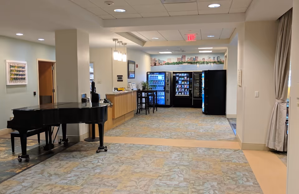 Interior view of a senior living facility hallway with a black grand piano on the left, a reception desk with hanging lights, vending machines at the end, and a cityscape mural on the back wall.