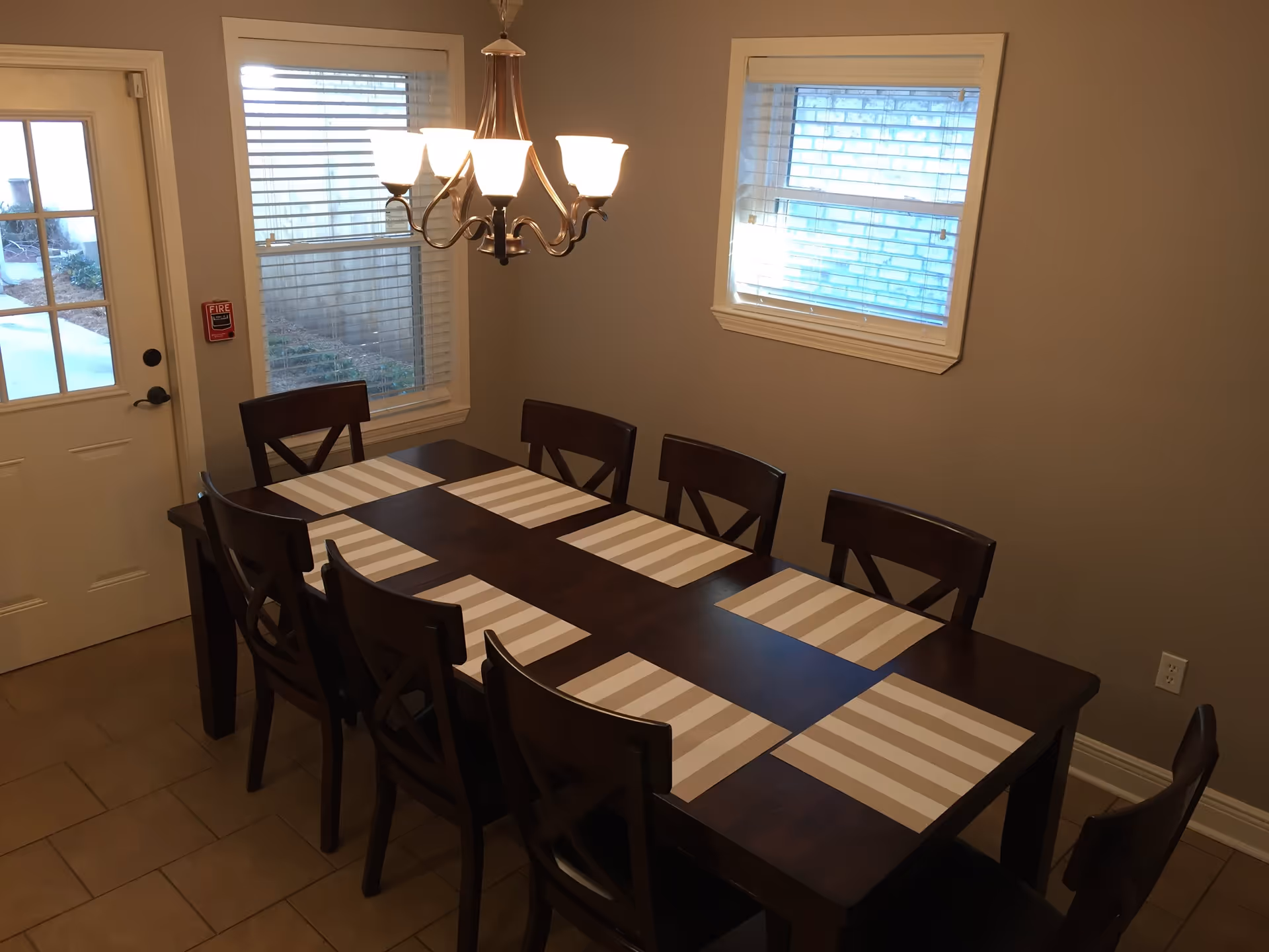 Dining room with a long dark wood table set with striped placemats, eight chairs, a chandelier, two windows, and a door.