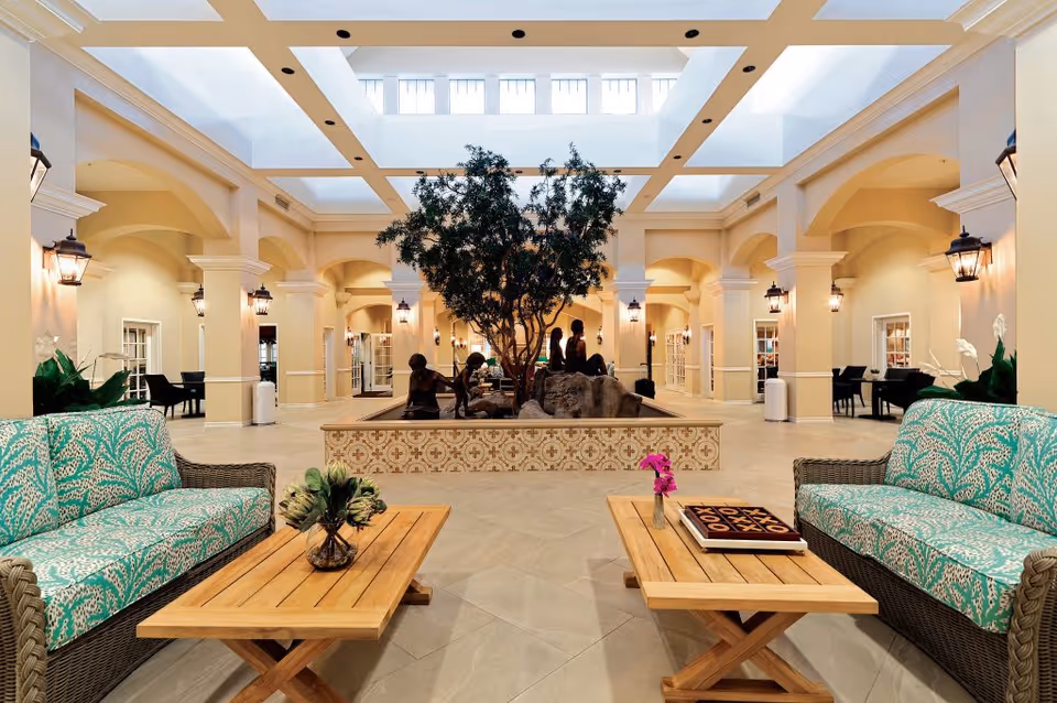 Spacious indoor atrium-style lounge with patterned wicker sofas, wooden coffee tables, and a central planter and sculpture beneath a skylight.