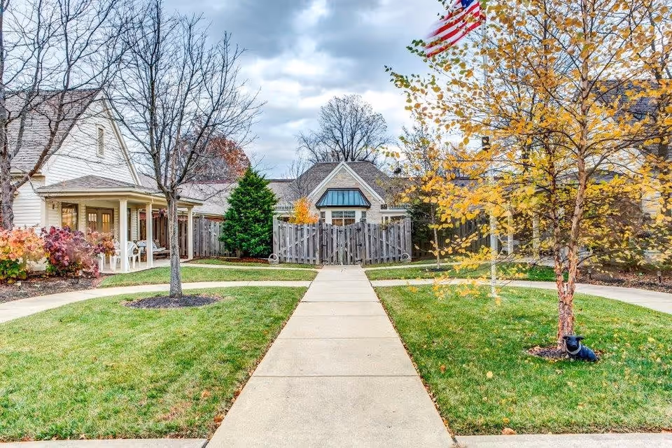 Concrete walkway through a landscaped courtyard leading to the front of townhome-style buildings with porches, a wooden fence, autumn trees, and an American flag.