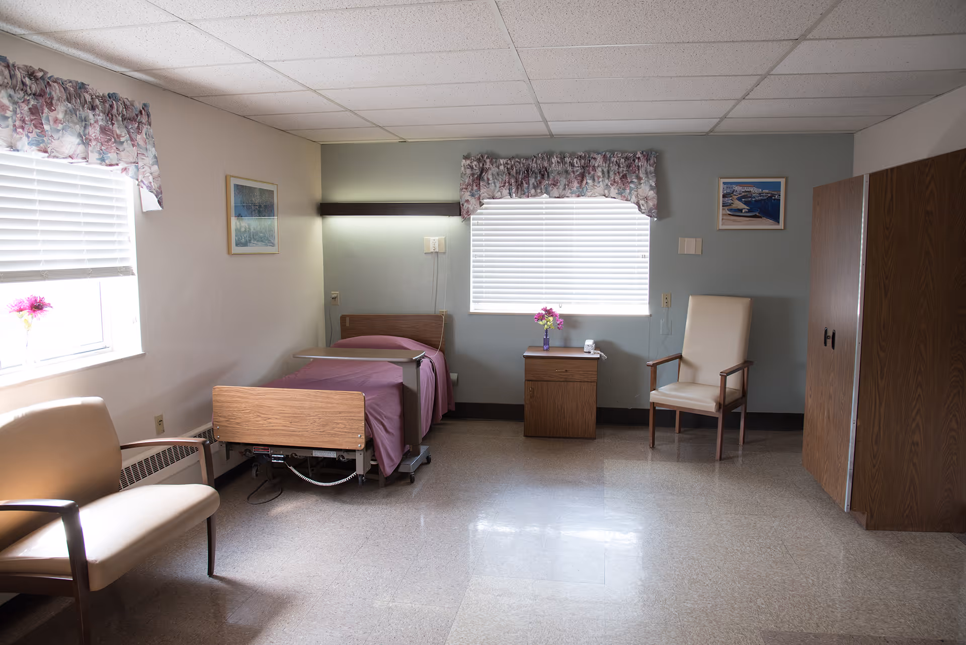 A simple, clean bedroom in a senior living facility with a single bed covered in a purple blanket, two chairs, a wooden nightstand with a vase of flowers, two windows with floral valances, and framed pictures on the walls.