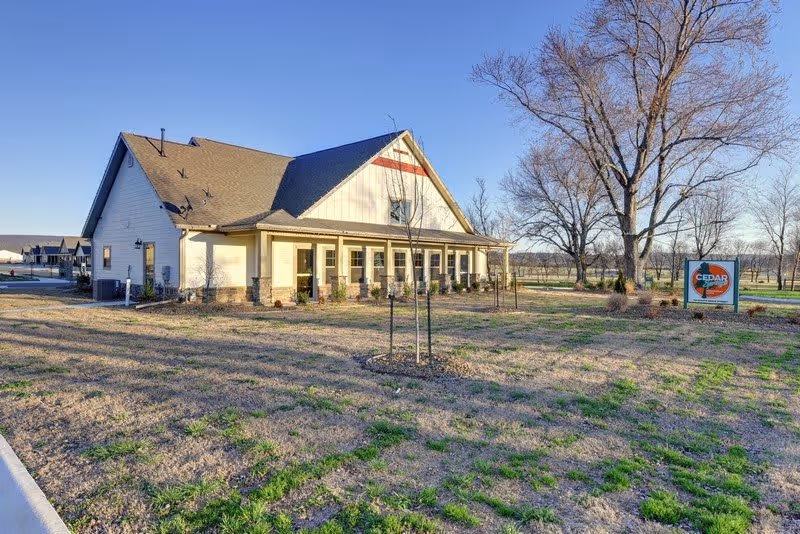 Exterior view of a single-story building with a pitched roof and a covered porch, surrounded by a grassy area with young trees and a sign that reads 'Cedar Springs Independent Living'. The sky is clear and the scene is well-lit by sunlight.
