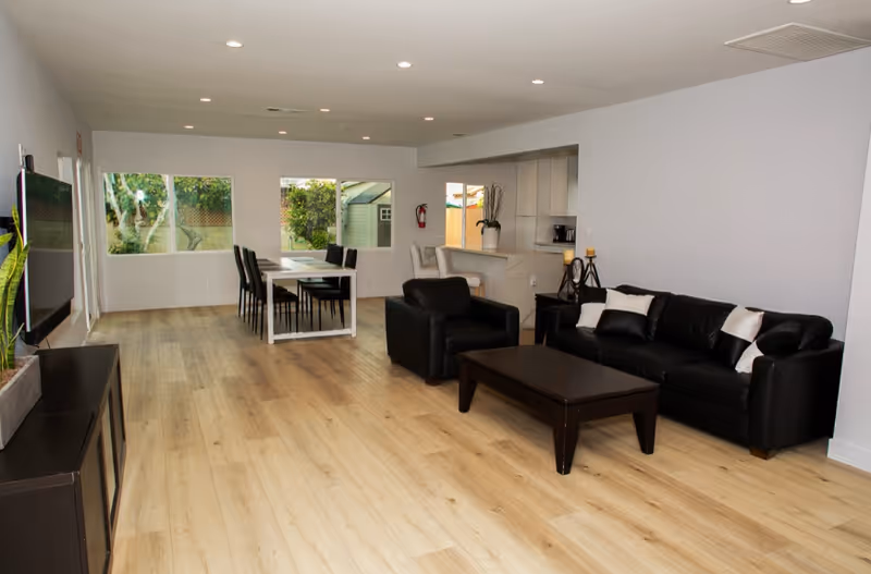 Spacious living area with black sofa set, coffee table, dining table, and large windows.