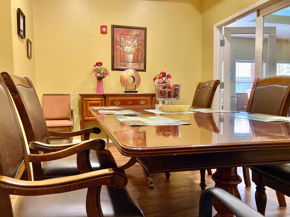 A polished wooden dining table with several chairs around it in a room with yellow walls. On the table are some papers and a glass bowl with decorative flowers. In the background, there is a wooden sideboard with two flower arrangements and a decorative sculpture, along with a framed floral painting on the wall. A doorway with glass doors leads to another room with windows.