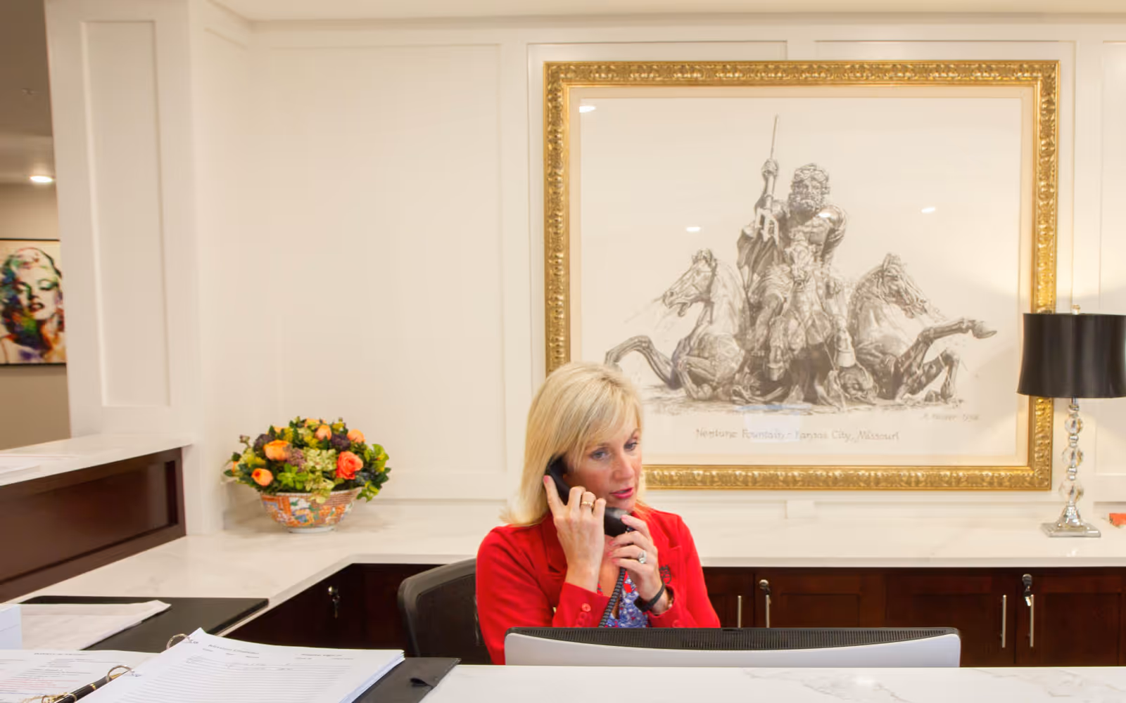A woman with blonde hair wearing a red jacket is sitting at a reception desk, talking on the phone. Behind her is a large framed artwork depicting Neptune Fountain in Kansas City, Missouri. The desk has a flower arrangement and a black lamp with a silver base.