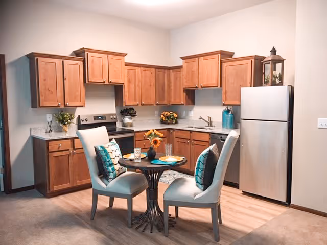 Open kitchen with wood cabinets, stainless refrigerator and stove, granite countertops, and a small round dining table with two upholstered chairs.