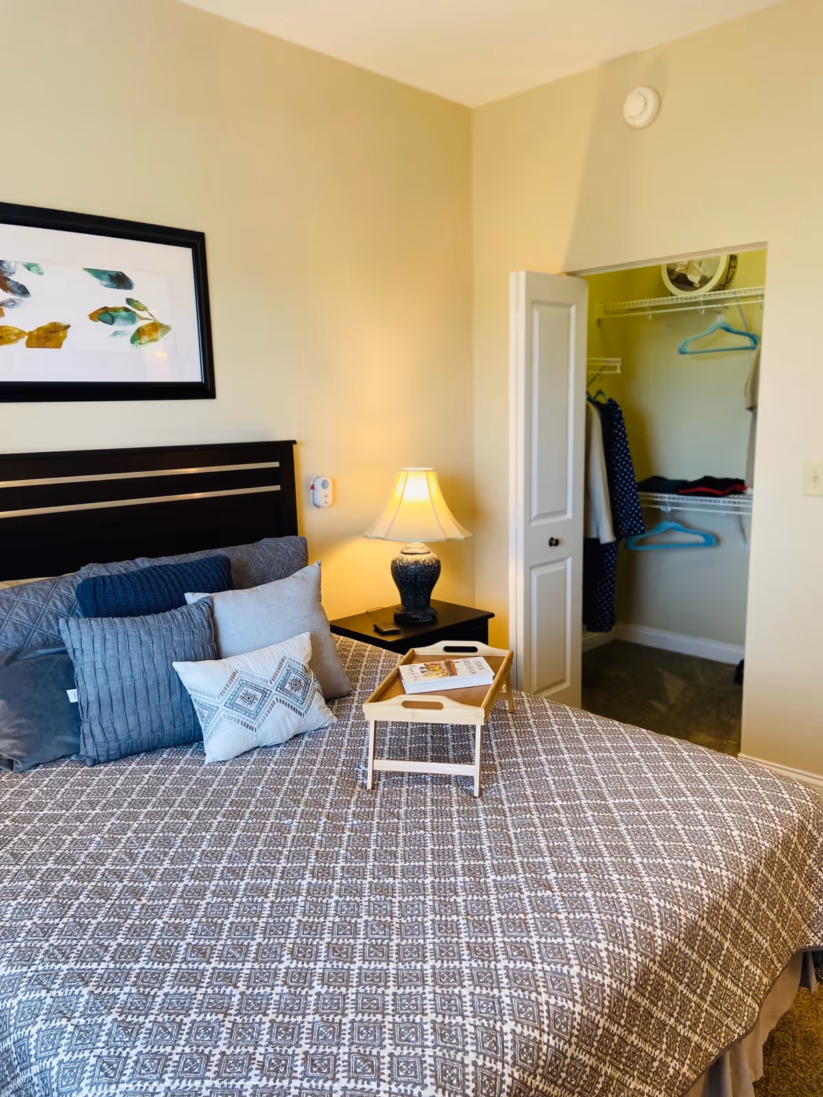 A bedroom with a neatly made bed featuring multiple pillows and a patterned bedspread. A small wooden tray with a book rests on the bed. To the right is a nightstand with a decorative lamp. An open closet with clothes hanging and shelves is visible in the background. The walls are painted light beige, and a framed abstract artwork hangs above the bed.