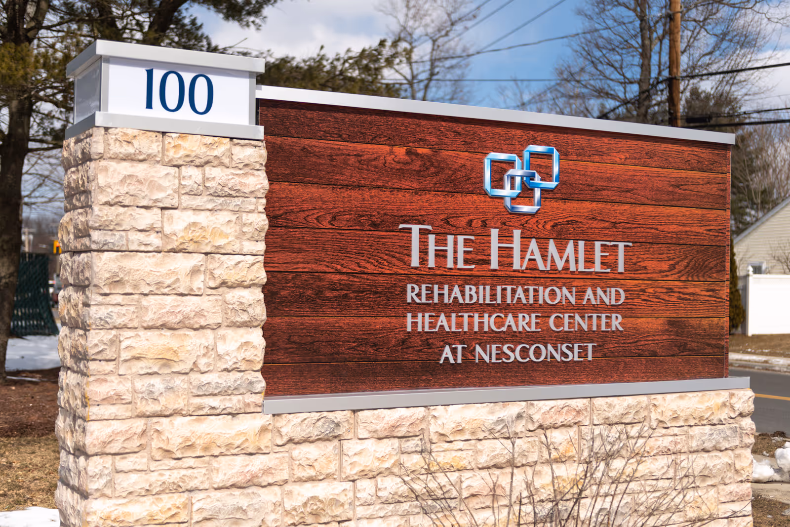 Outdoor stone and wood sign for The Hamlet Rehabilitation and Healthcare Center at Nesconset with the number 100 on a white panel at the top left, set against a background of trees and a partly cloudy sky.