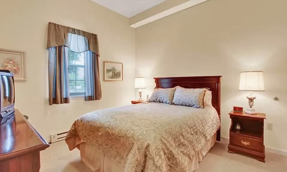 A cozy bedroom with a large bed covered in a beige patterned bedspread and matching pillows. There are two wooden nightstands on either side of the bed, each with a lamp. A window with blue and brown curtains is on the left wall, and two framed pictures hang on the walls. A wooden dresser with a TV on top is visible on the left side of the room.