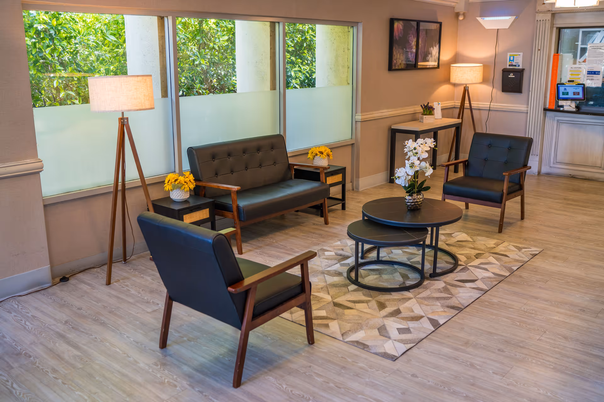 A cozy seating area in a senior living facility with two black armchairs and a black loveseat arranged around two nested round coffee tables on a patterned rug. The room has light wood flooring, two floor lamps with beige shades, small side tables with yellow flowers, and large windows with frosted lower panes letting in natural light. A reception desk is visible in the background.