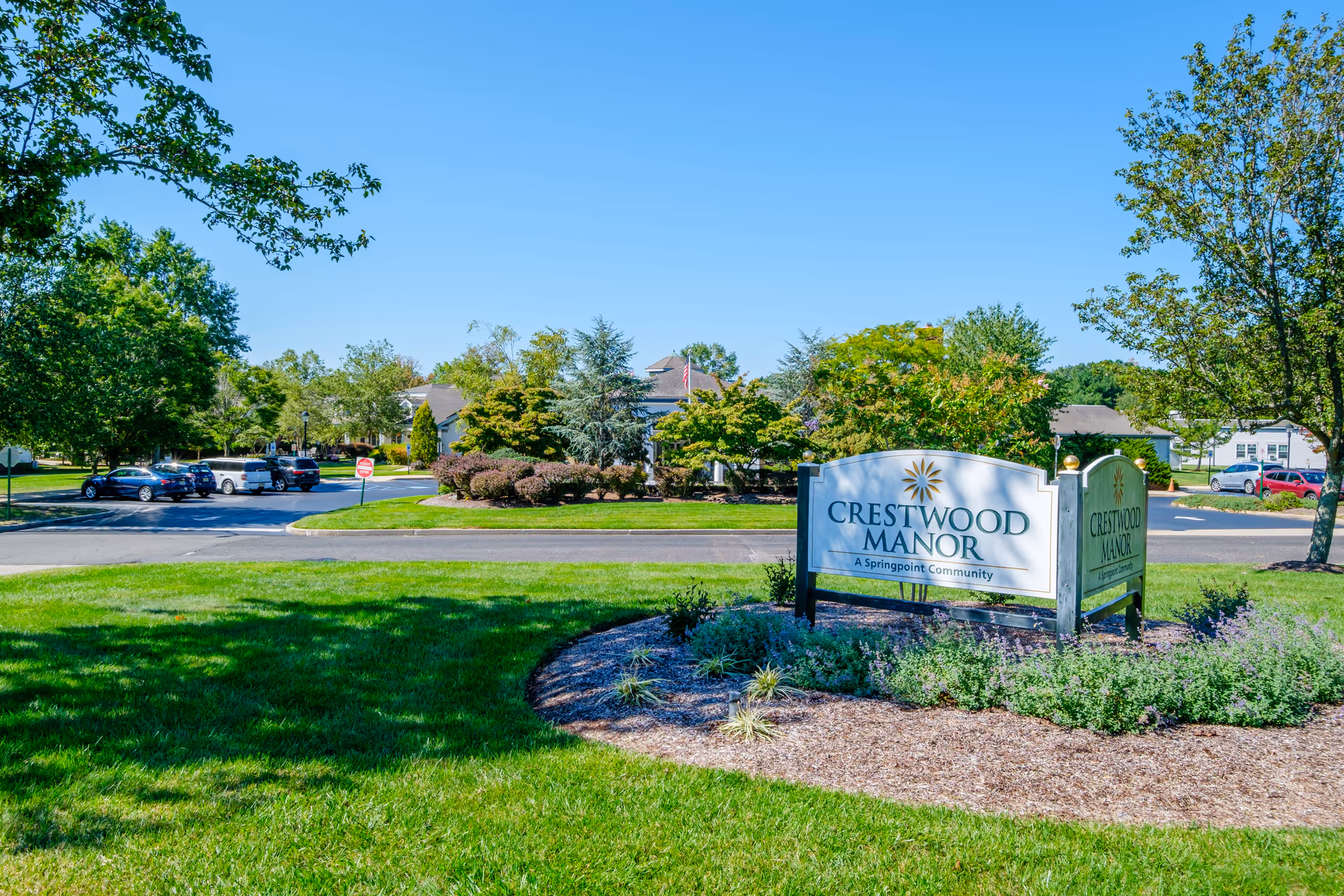 Outdoor view of Crestwood Manor senior living facility entrance sign surrounded by landscaped greenery and trees, with parked cars and buildings visible in the background under a clear blue sky.