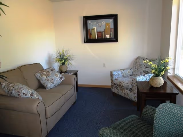 A small living room with a beige couch adorned with patterned pillows, a patterned armchair, a green armchair, two wooden side tables each holding a vase with yellow flowers, a framed abstract painting on the wall, and a window letting in natural light.