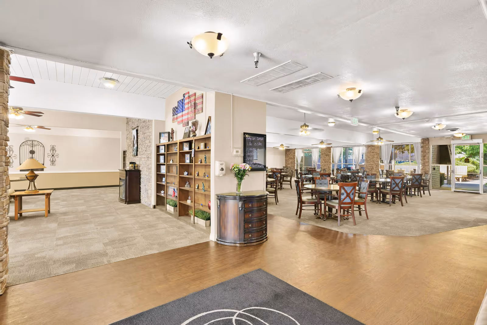 Interior view of a senior living facility showing a spacious dining area with multiple tables and chairs. The room has large windows with white curtains allowing natural light to enter. There is a wooden bookshelf with decorative items and an American flag on the wall. The flooring transitions from wood near the entrance to carpet in the dining area. Ceiling lights and fans are visible, and an exit door leads outside to a garden or patio area.