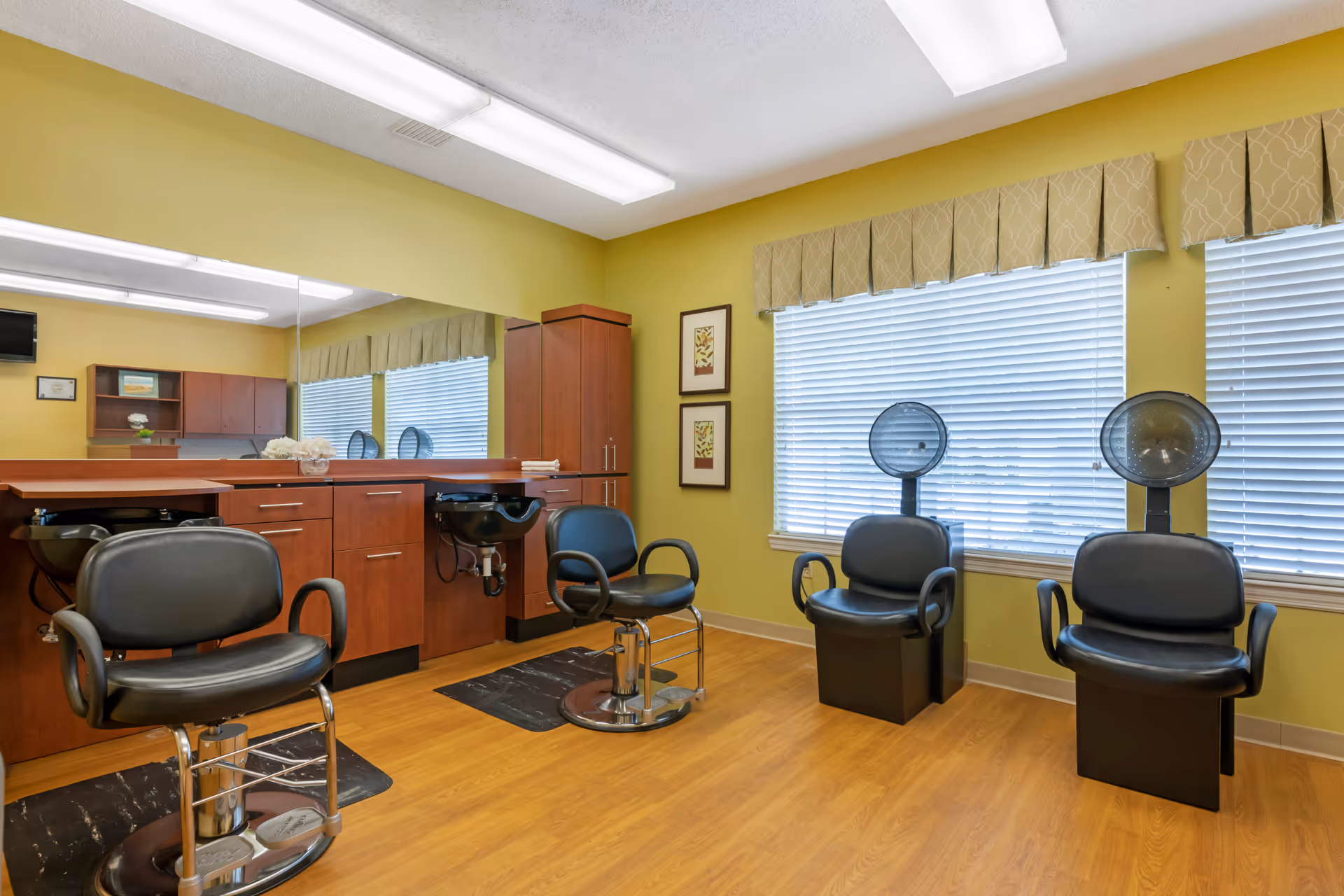 A bright salon area with styling chairs, hooded hair dryers, sinks and wooden cabinetry against yellow-green walls and large windows.