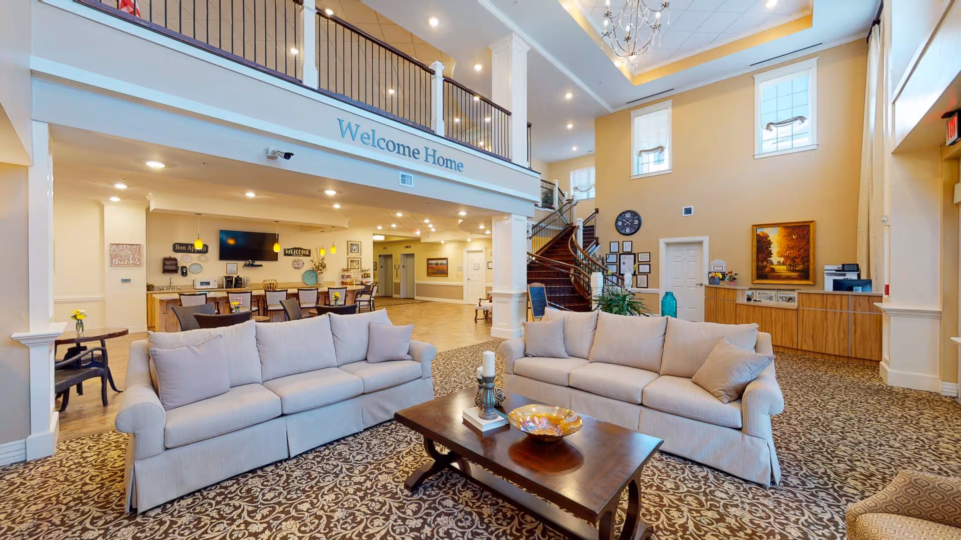 A spacious and well-lit living room area in a senior living facility with two beige sofas facing each other, a wooden coffee table with decorative items, a patterned carpet, and a high ceiling with large windows. In the background, there is a reception desk, a staircase leading to an upper floor, and a sign that reads 'Welcome Home'.