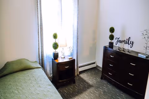 Bedroom in a senior facility with bed, nightstand lamp, dresser, and 'Family' sign.