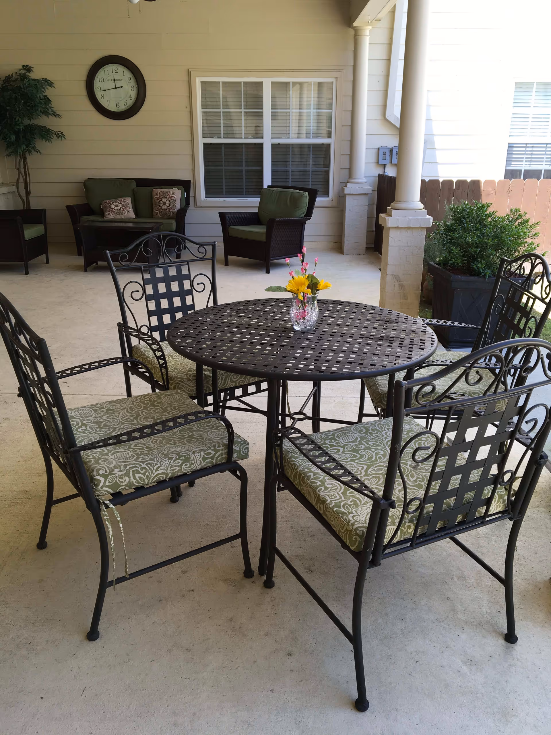 Outdoor covered patio area with a round metal table and four metal chairs with green patterned cushions. A small vase with yellow and pink flowers is on the table. In the background, there are two armchairs with green cushions and a small table, a wall clock, a window, and some potted plants.