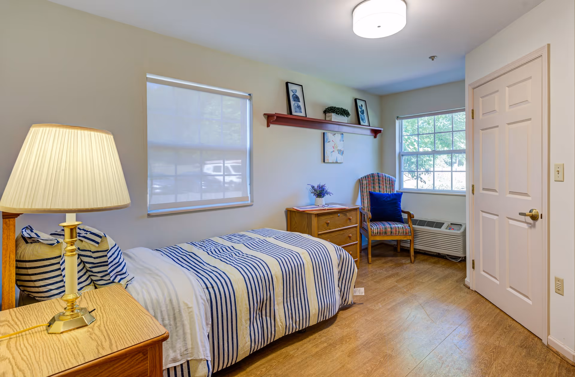 A small, neatly arranged bedroom with a single bed covered in white and blue striped bedding. A wooden nightstand with a lamp is beside the bed. There is a wooden dresser with a small plant and a book on top, a striped armchair with a blue cushion, and two windows with white blinds letting in natural light. The walls are light-colored, and there is a red shelf with framed pictures above the dresser. The floor is wooden, and a closed white door is visible on the right side.
