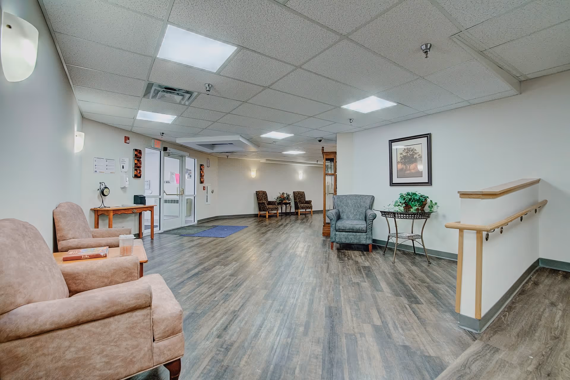 A spacious and well-lit interior common area with wooden flooring, beige and gray upholstered chairs, a small wooden table with a clock and books, a framed picture on the wall, and a side table with a plant. The area has white walls, ceiling lights, and a handrail along a short staircase.