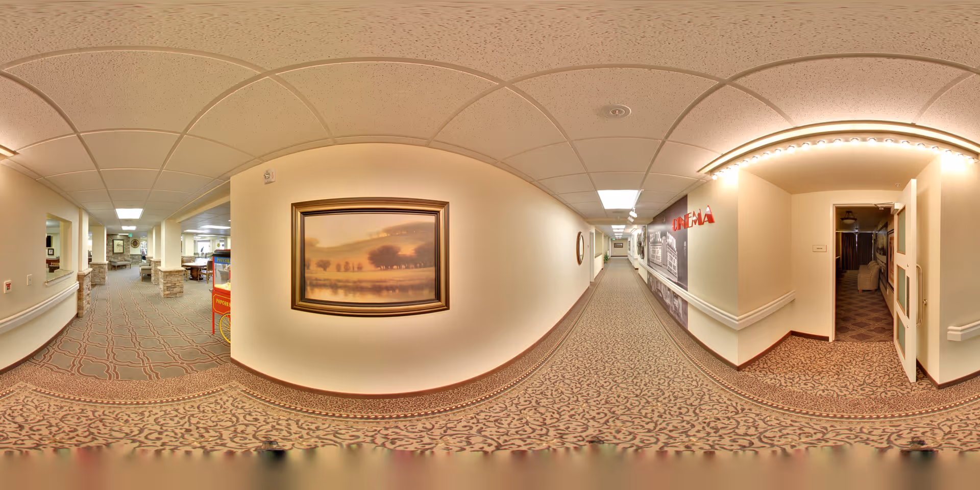 A wide hallway in a senior living facility with patterned carpet and beige walls. On the left side, there is an open area with stone pillars and seating. On the right side, there is a door labeled 'Cinema' leading to a small theater room. The hallway is well-lit with ceiling lights and has a framed landscape painting on the wall.