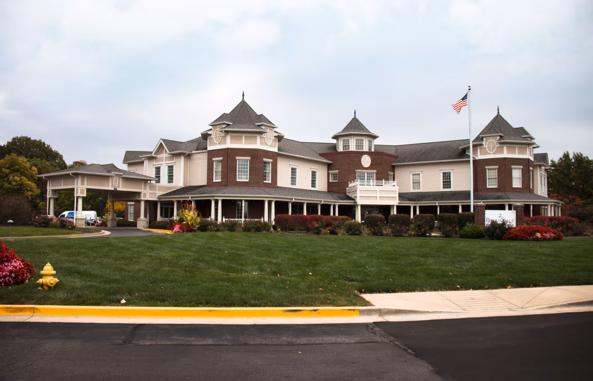 Front view of a large two-story senior living facility with turrets, a covered entrance, manicured lawn, and an American flag.