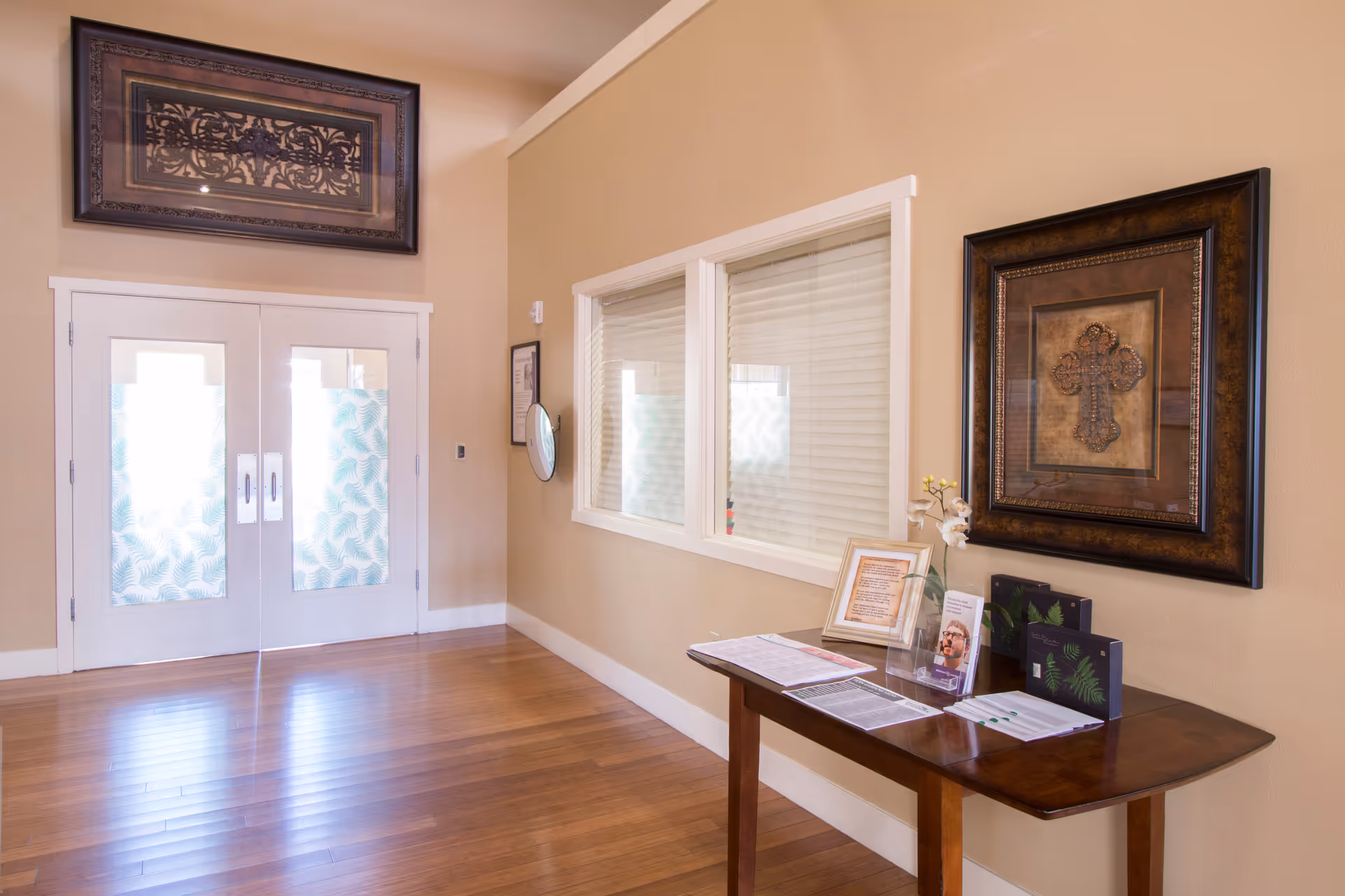 Interior hallway with wooden flooring and beige walls featuring a double door with frosted glass panels, a wooden table with brochures and framed documents, and two framed artworks on the walls.