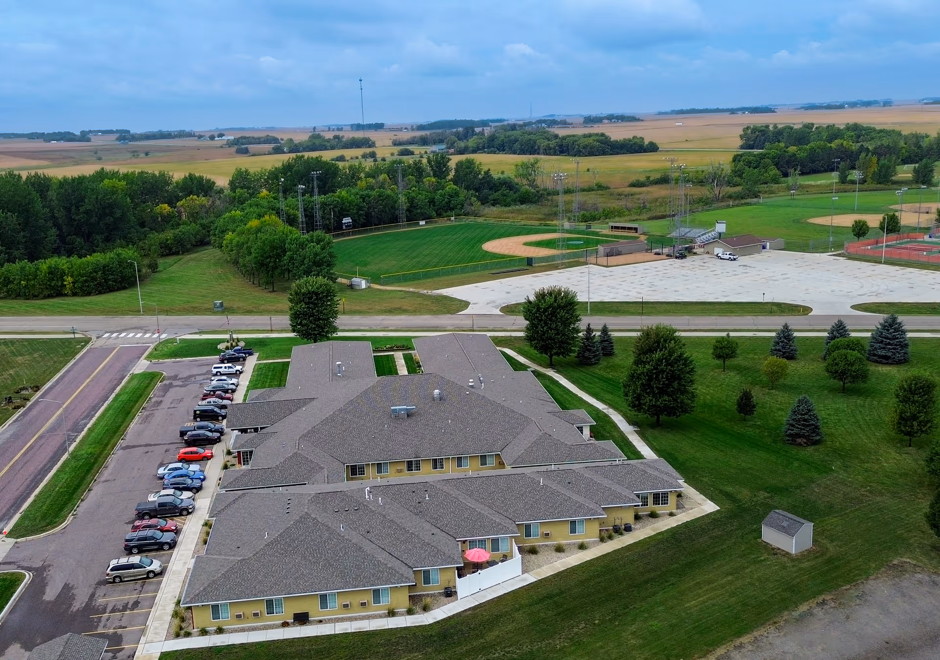 Aerial view of a large single-story building with a gray roof and yellow walls, surrounded by green lawns and trees. There is a parking lot with several cars parked along one side of the building. In the background, there are baseball fields and open farmland under a cloudy sky.