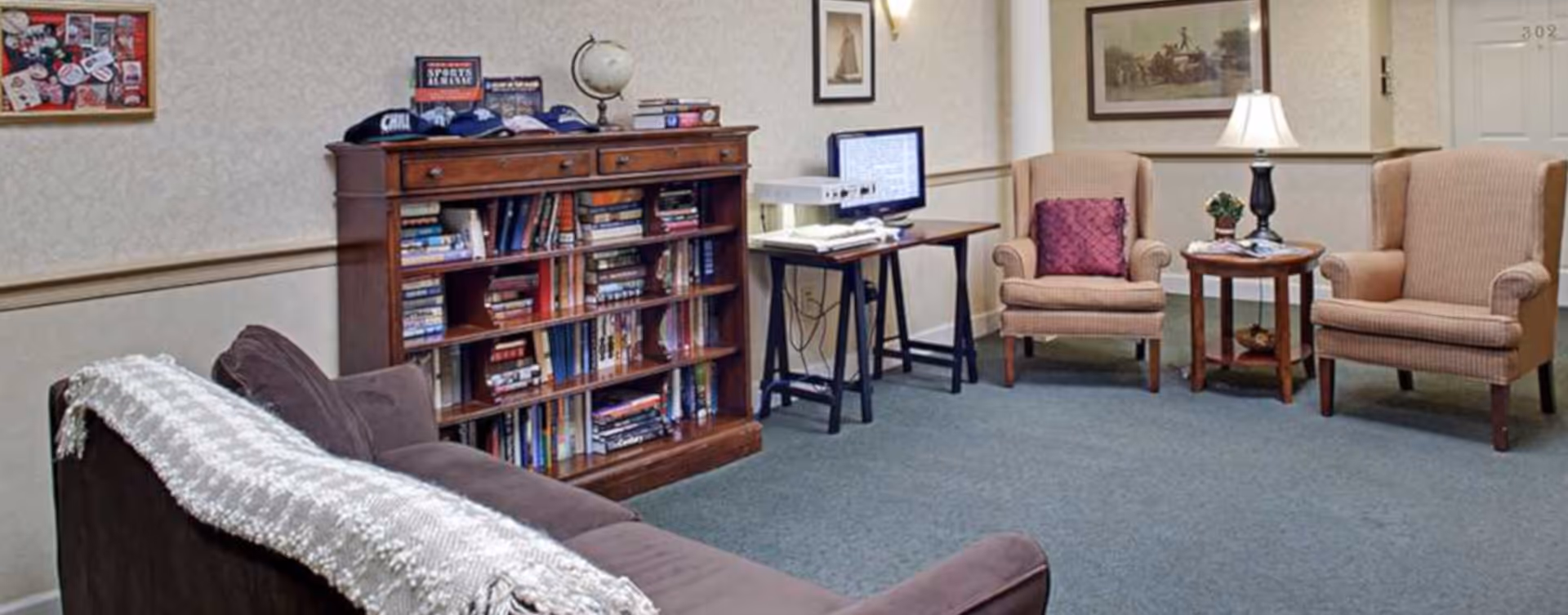 Comfortable sitting area with a bookshelf, computer desk, sofa and two armchairs in a senior living facility.