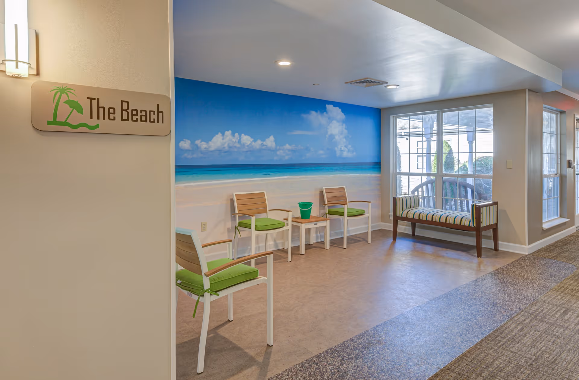 Indoor seating area named 'The Beach' with a wall mural depicting a beach scene with blue sky, clouds, and ocean. The area has three chairs with green cushions, a small white table with a green bucket on it, and a striped cushioned bench near large windows letting in natural light.