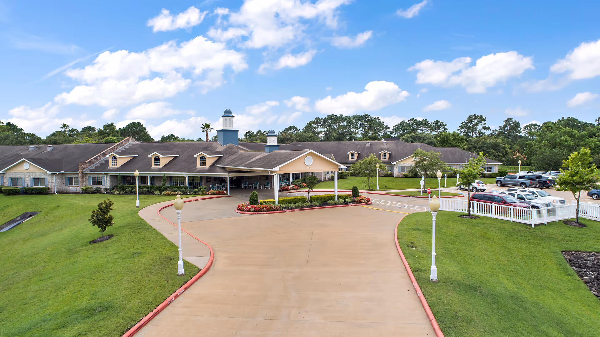 Front entrance and circular driveway of a single-story senior living facility with landscaped lawns and parked cars.