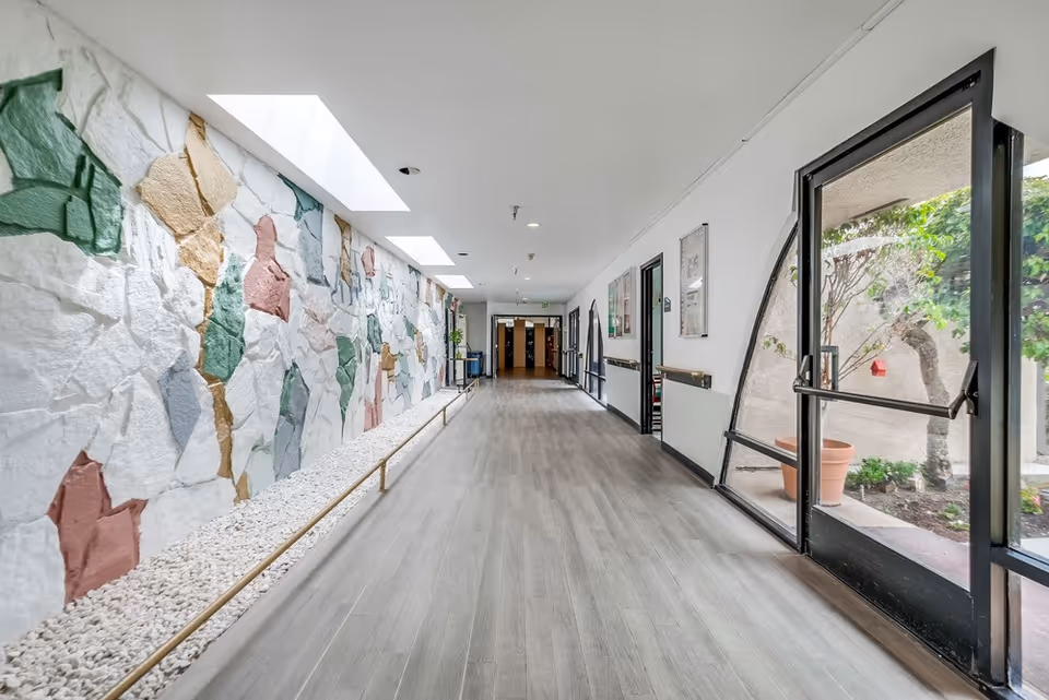 A long, bright hallway in a healthcare facility with a colorful stone accent wall on the left and large glass doors on the right leading to an outdoor garden area. The floor is wood-style laminate, and there are skylights in the ceiling allowing natural light to illuminate the space.