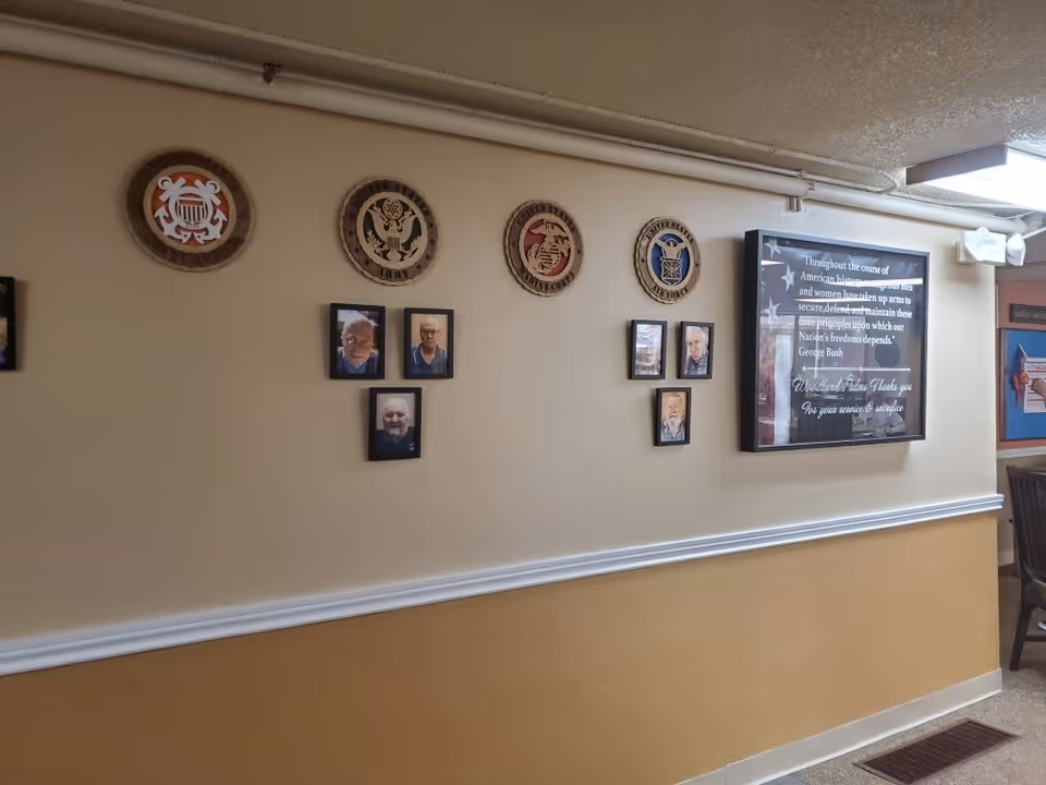 Interior hallway wall at Woodland Palms Memory Care decorated with framed photos of elderly individuals and circular plaques representing the United States Coast Guard, Army, Marine Corps, and Air Force. A framed quote from George Bush thanking veterans is also displayed on the wall.