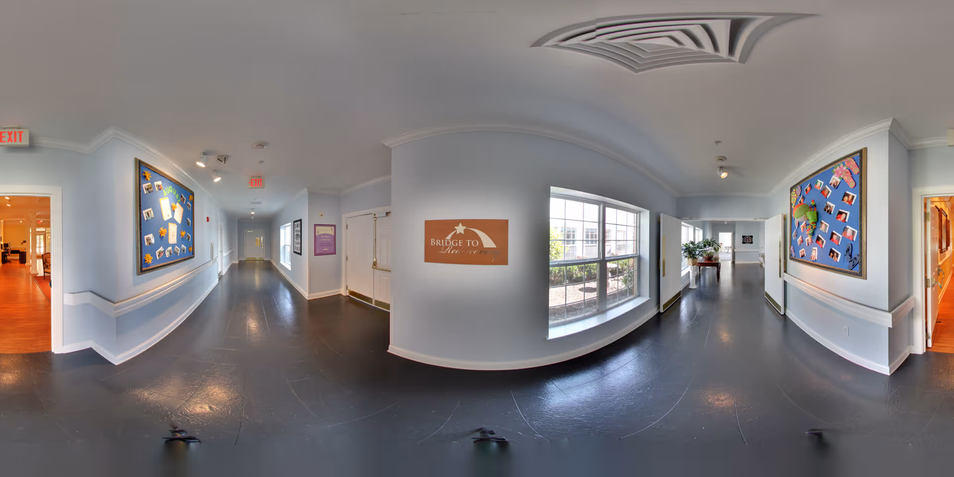 A wide hallway in a senior living facility with light blue walls and dark flooring. The hallway features bulletin boards with photos and notes on both sides, a large window letting in natural light, and exit signs above doorways. There are handrails along the walls and a sign that reads 'Bridge to New Life' mounted on the wall near the window. The hallway leads to other rooms with wooden floors visible through open doorways.