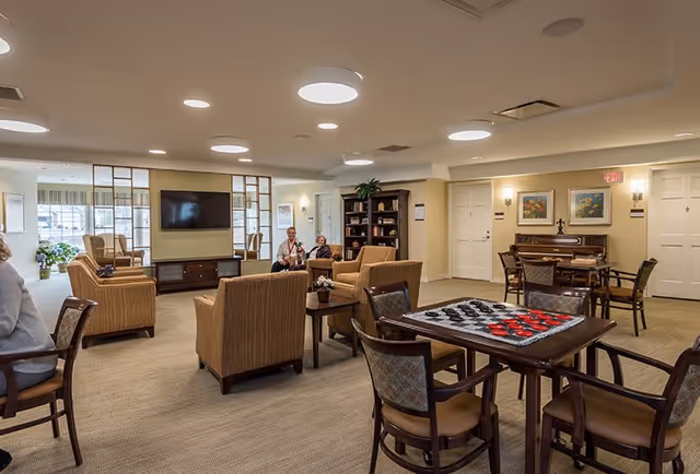 A spacious and well-lit common area in a senior living facility featuring comfortable armchairs arranged around a coffee table, a large flat-screen TV mounted on the wall, a bookshelf, and a piano with chairs. A table in the foreground has a checkers game set up. Two people are seated and conversing near the center of the room.