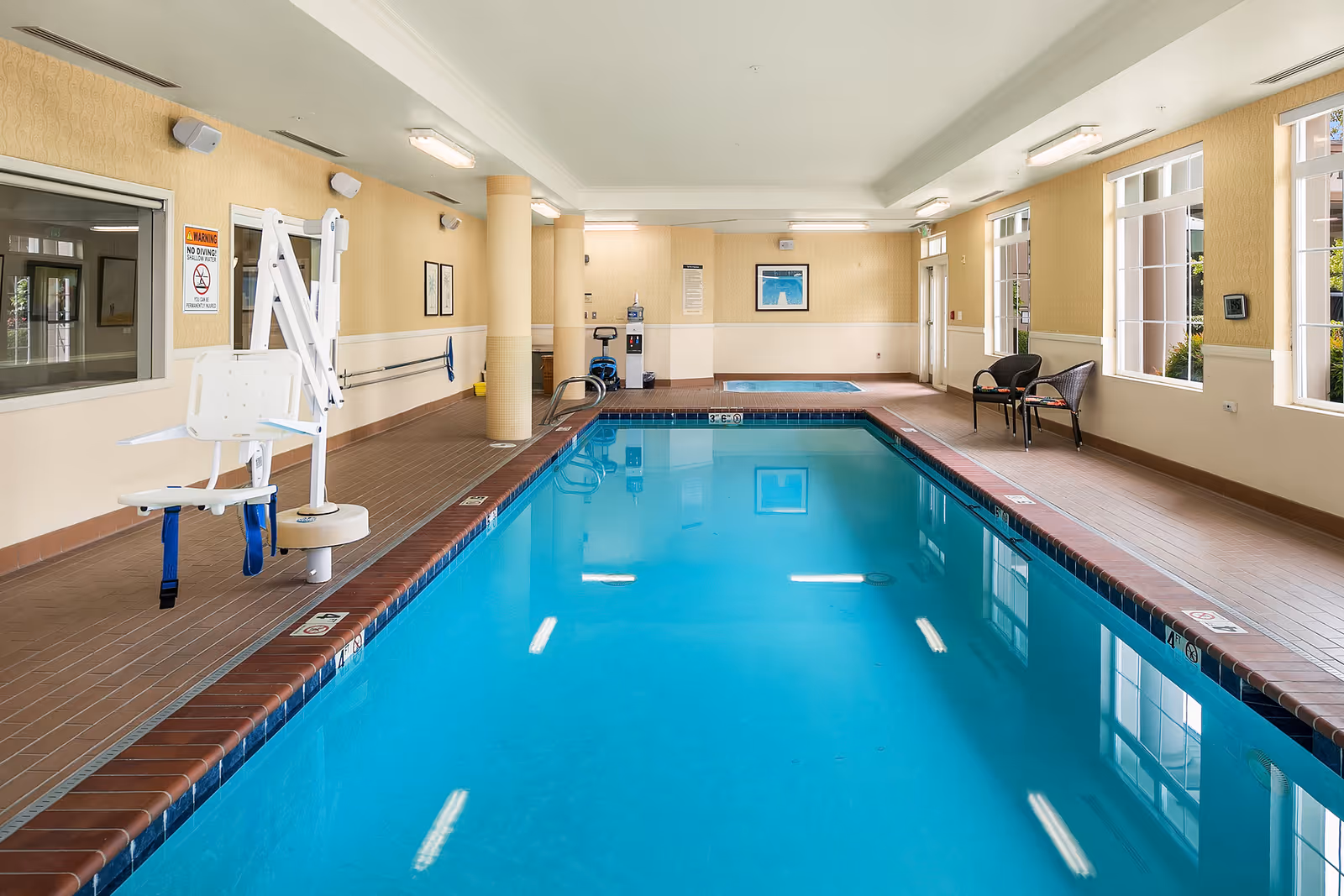 Indoor swimming pool with clear blue water, surrounded by beige walls and large windows letting in natural light. There is a pool lift chair on the left side for accessibility, two black chairs on the right side near the windows, and a blue mat at the far end of the pool area.