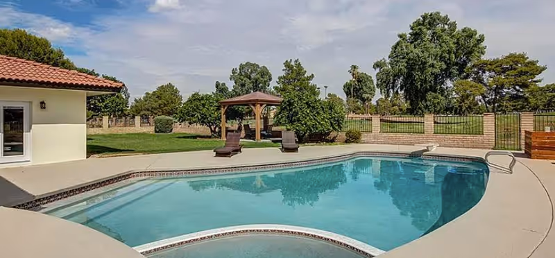 Outdoor swimming pool with clear blue water surrounded by a concrete deck. Two lounge chairs and a wooden gazebo are visible on the grassy area next to the pool. Trees and a brick fence enclose the area under a partly cloudy sky.